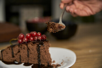 Tasting a slice of chocolate cake with a fork. Homemade dark chocolate dessert is fresh from the oven and decorated with melted chocolate glaze, powdered white sugar, red currants and blueberries
