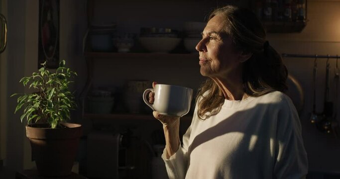 Cinematic shot of happy smiling senior pensioner woman is drinking hot tea or american coffee and looking through window with sunlight in morning in kitchen at home.Concept of comfort, family, elderly