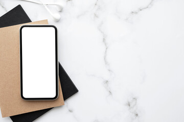 Smartphone with white blank mock up screen is on top of white marble desk table. Top view with copy...