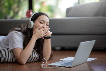 Asian woman wearing headphones is sleepy  studying online using a laptop at home.