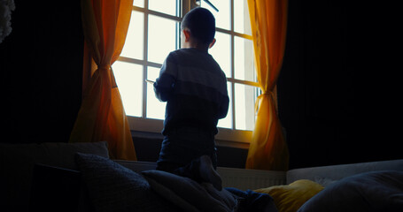Little lonely boy looks out the window and holds a tablet in his hand, a child in isolation, boy in...