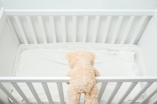 Brown Teddy Bear On Railing Of White Crib. Concept Of Baby Safety Climbing In Bed. Back View. Top Down View.