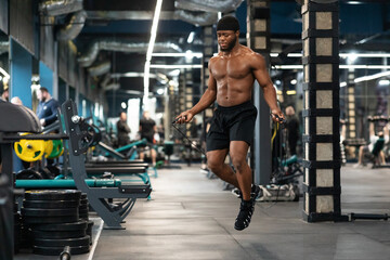 Fototapeta na wymiar Shirtless muscular african man jumping with skipping rope in gym