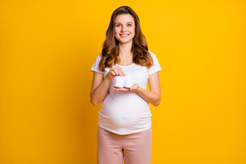 Portrait of attractive careful cheerful pregnant girl holding in hands knitted socks unborn isolated over bright yellow color background