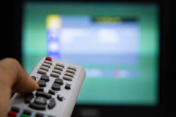 Man's hand holding TV remote control with a television in the background.