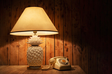 Antique table lamp with old phone on the wooden table in front of wooden wall in the room.
