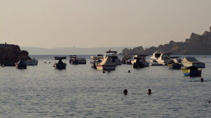 Zatoka z jachtami ze skalistym brzegiem, daleko w tle widać sylwetkę wyspy Gozo, Malta  - obrazy, fototapety, plakaty