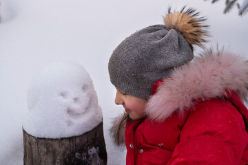 A girl in a winter hat and a red jacket with a small snowman in the snow. The concept of winter fun