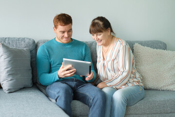 son teaching his mother to use tablet. older people using technology. Cheerful elderly woman sitting on the sofa next to his adult son