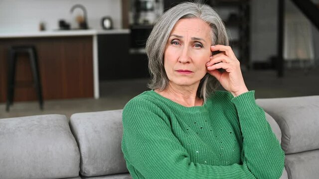 Thoughtful sad ged woman with grey hair, thinking about the future sitting on the sofa at apartment, planning life, worried about her children, home and property, inability to pay bills