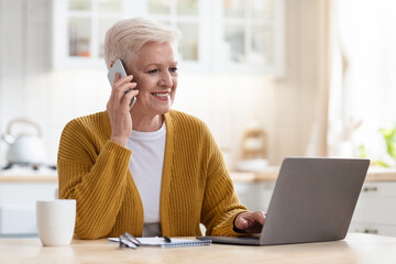 Smiling senior woman having conversation on phone, working on laptop