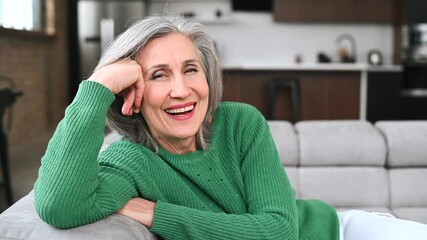 Joyful senior mature full of energy woman in casual green sweater resting on the sofa with hands behind head, sitting on the couch in modern living room, middle-aged lady posing and looking at camera