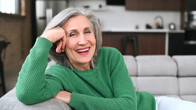Joyful senior mature full of energy woman in casual green sweater resting on the sofa with hands behind head, sitting on the couch in modern living room, middle-aged lady posing and looking at camera