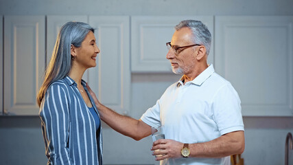 Obraz premium side view of smiling senior man holding glass of water while touching shoulder of happy asian wife