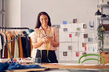 Female Fashion Designer Browsing Internet On Mobile Phone Stands In Studio With Measure Around Neck