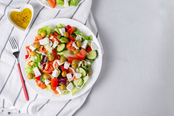 Delicious vegetable greek salad with feta, tomatoes, olives, greens on light background. Served with towel, fork and olive oil dressing. Healthy diet lunch idea. Quick, simple recipe.