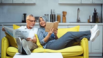 Fototapeta premium cheerful asian woman showing book to elderly husband holding newspaper