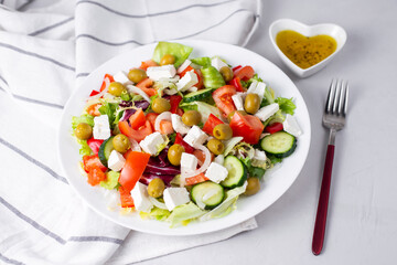 Delicious vegetable greek salad with feta, tomatoes, olives, greens on light background. Served with towel, fork and olive oil dressing. Healthy diet lunch idea. Quick, simple recipe.