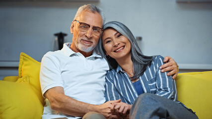 cheerful elderly interracial couple smiling at camera while hugging on sofa at home