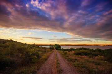 Fototapeta na wymiar Rural road leading to the lake, river, at sunset, against the backdrop of beautiful clouds and the evening sky