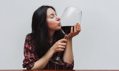 A young girl in a plaid shirt with closed eyes tenderly holds a large glass of red wine and kisses it on a gray background.Concept of love for alcohol, alcoholism, hangover, loneliness and depression.