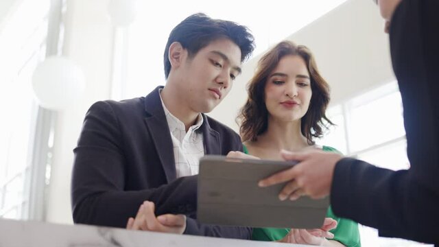 Multiethnic couple contact make documents to stay in hotel. Reception counter provide accommodation booking service through tablet. Group businessmen meeting plan to work. Concept Residential business