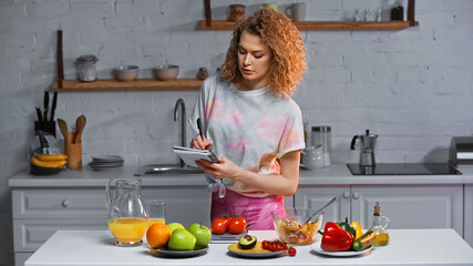 woman writing in notebook and weighing tomatoes near vegetables and orange juice on table