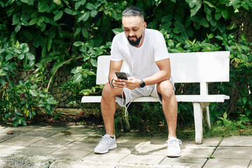 Positive handsome bearded man resting on bench in park, listening to music and texting friends