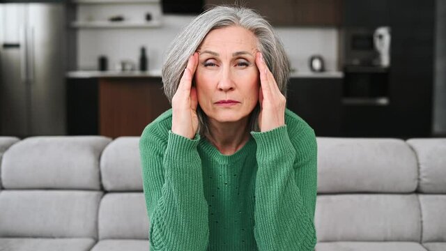 Frustrated senior woman feels headache, migraine, a mature retired lady sits on the couch at home and holds forehead, a grandmother has memory losses, suffers from pain in the temples