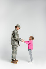 Side view of smiling soldier and daughter holding hands on grey background