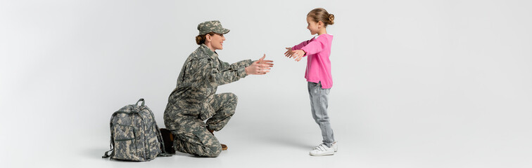 Fototapeta premium Side view of soldier outstretching hands near smiling daughter on grey background, banner