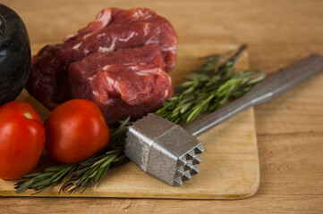A large piece of pork with meat hammer and two sprigs of rosemary on a wooden table. Ingredients for cooking meat dishes.