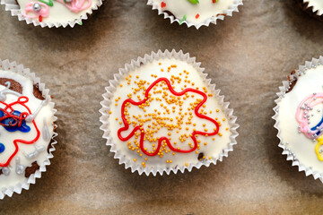 Chocolate brown muffins wrapped in white paper and covered with white frosting with colorful decorations, baked in the oven, lying on baking paper, top view.