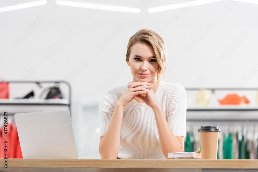 Wall mural Young seller looking at camera near laptop, notebook and coffee to go in showroom