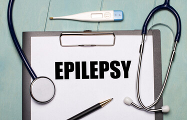 On a light blue wooden background, there is a paper labeled EPILEPSY, a stethoscope, an electronic...