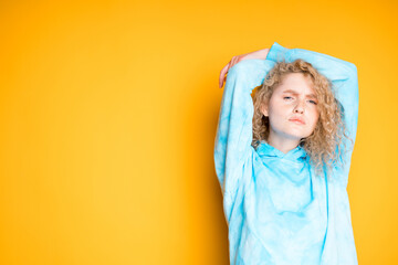 A young girl in a blue sweater raises her hands up behind her head on a bright background