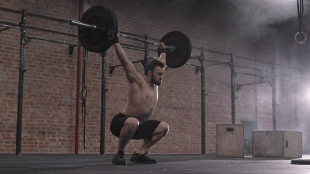 Wide shot of young handsome Caucasian sportsman with naked torso lifting up heavy barbell in gym and then throwing it on the floor