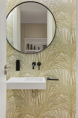 Bathroom with wallpaper with plant leaves texture, small wash basin and round mirror. Minimalist interior