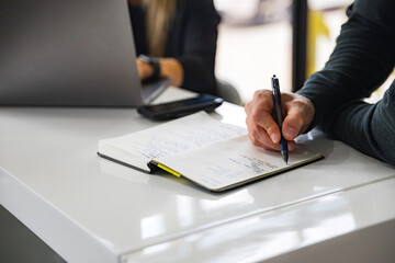 Man writing in notebook next to coworker on laptop and phone in the morning office meeting with a...