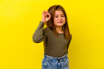 Little caucasian girl isolated on yellow background winks an eye and holds an okay gesture with hand.