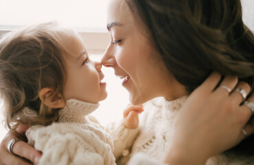 Young mother hugging her little daughter, they sitting near window and smiling. 