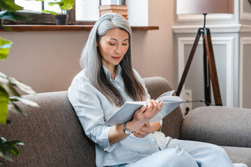 Pretty caucasian mature woman sitting on the sofa and reading a book