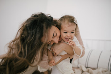 Young mother and her little daughter having fun in a bedroom, they playing, laughing and hugging. Happy family concept.