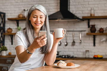 Happy middle-aged woman using smart phone during the breakfast in the kitchen