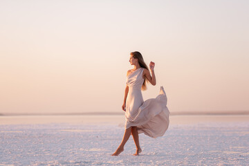 Young blonde woman in an evening airy pastel pink, powdery dress stands barefoot on white...