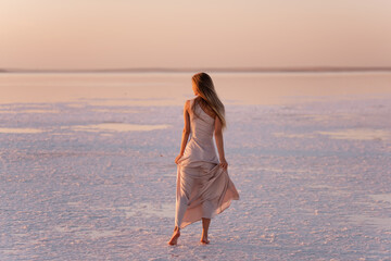 Young blonde woman in an evening airy pastel pink, powdery dress stands barefoot on white crystallized salt. Girl with natural make-up, hair is developing. Salt mining trip, walking on water at sunset