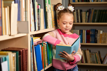 child girl get carried away by reading a book in library, standing betweel multi coloured...