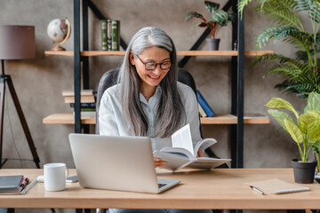 Matured caucasian grey-haired woman reading a book at office desk