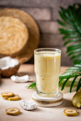 banana cocktail with coconut milk in a glass on light beige background with palm leaves