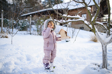 Small girl outdoors in winter garden, standing by wooden bird feeder.